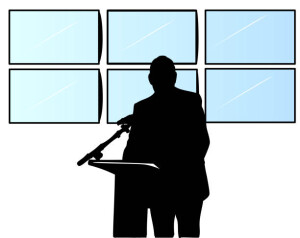 Public speaker in silhouette with television screens behind him